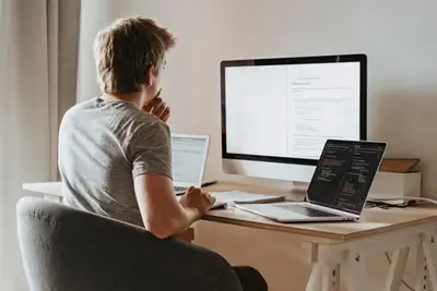 A software engineer at a desk, reading code on a monitor and a laptop